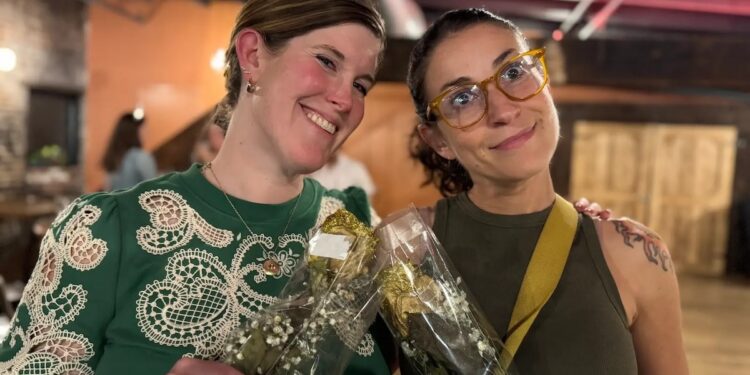 Two women stand together indoors, smiling and posing for a photo. The woman on the left has shoulder-length brown hair and is wearing a green lace blouse and gold earrings. The woman on the right has her dark hair tied back, wears yellow-rimmed glasses, and has a tattoo on her left shoulder. They are both holding small bouquets of white baby’s breath wrapped in clear plastic. The background is a dimly lit room with industrial-style ceilings.