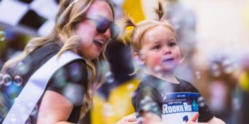 A close-up of a young girl with two pigtails and a "#1" racing bib, smiling as she stands among floating bubbles; behind her, a woman wearing a tiara and sunglasses looks on with a happy expression.