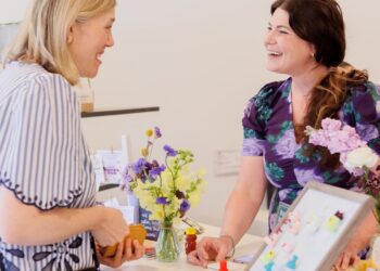 A woman with blonde hair in a striped blue and white shirt stands at a white counter, smiling and talking to a brunette woman in a purple floral dress. The woman in the floral dress is behind the counter, which displays small crafts, a vase of yellow and purple flowers, and a framed display of colorful pins or magnets.
