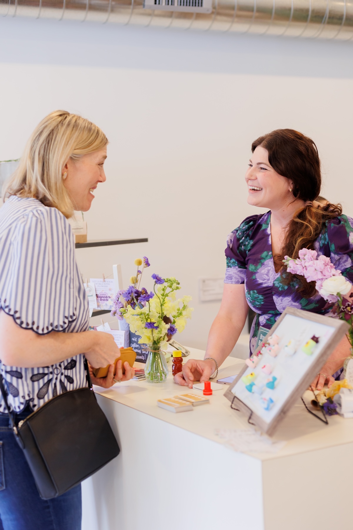 A woman with blonde hair in a striped blue and white shirt stands at a white counter, smiling and talking to a brunette woman in a purple floral dress. The woman in the floral dress is behind the counter, which displays small crafts, a vase of yellow and purple flowers, and a framed display of colorful pins or magnets.