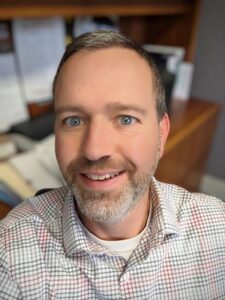A close-up professional headshot of a man with light-colored eyes and short, graying hair and a trimmed beard. He is smiling warmly at the camera. He is wearing a white, button-down dress shirt with a fine grid pattern of red and gray lines. The background is a slightly blurred office setting, with a wooden desk or shelf visible.