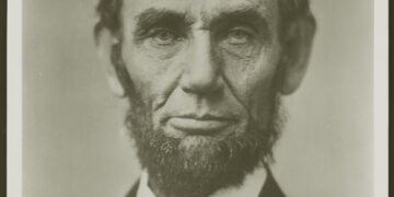 A detailed, head-and-shoulders black-and-white portrait of a beardless Abraham Lincoln, photographed by Alexander Hesler in Springfield, Illinois, on June 3, 1860. Lincoln is facing slightly right, with a thoughtful and calm expression. He has dark, somewhat tousled hair, deep-set eyes, and visible lines etched into his face. He is wearing a dark suit jacket, a white collared shirt, and a dark, slightly askew bowtie. The background is a plain, light grey tone.