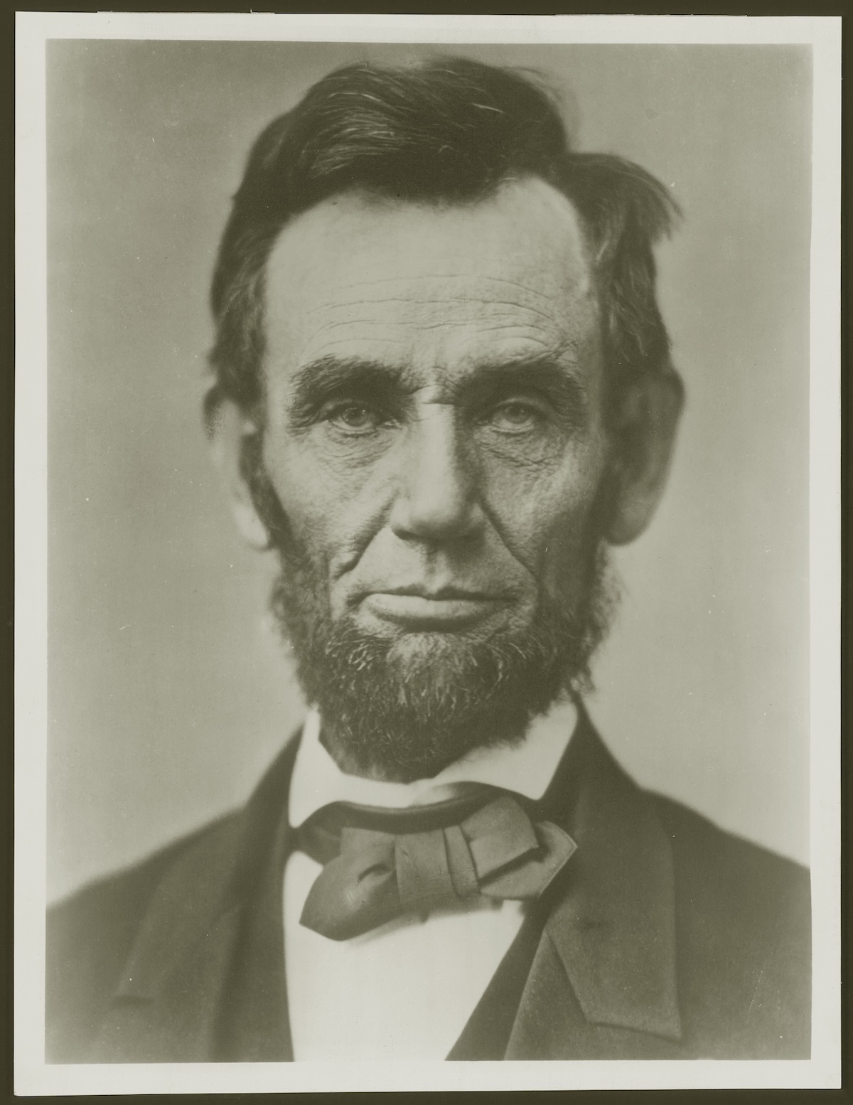 A detailed, head-and-shoulders black-and-white portrait of a beardless Abraham Lincoln, photographed by Alexander Hesler in Springfield, Illinois, on June 3, 1860. Lincoln is facing slightly right, with a thoughtful and calm expression. He has dark, somewhat tousled hair, deep-set eyes, and visible lines etched into his face. He is wearing a dark suit jacket, a white collared shirt, and a dark, slightly askew bowtie. The background is a plain, light grey tone.