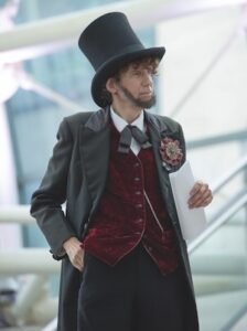 A medium shot of a professional Abraham Lincoln impersonator, likely Danny Russel, standing in a modern indoor space. He is dressed in an authentic 19th-century costume, featuring a tall black silk top hat, a long dark frock coat, and a distinctive deep red velvet vest. A large, multi-colored floral rosette pin is attached to his lapel, and a silver pocket watch chain is visible across his vest. He holds a white document in his hand, appearing to be in character for a performance or presentation.