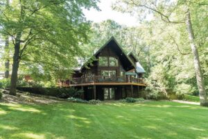 A large, multi-story log cabin with a steep gabled roof and expansive glass windows is nestled among mature, leafy green trees. A wide wooden deck with a dark railing wraps around the main level, overlooking a sprawling, neatly manicured green lawn. The lower level features glass doors leading out to a small stone patio. The scene is bright and airy, captured on a sunny day with soft shadows dappling the grass.