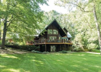 A large, multi-story log cabin with a steep gabled roof and expansive glass windows is nestled among mature, leafy green trees. A wide wooden deck with a dark railing wraps around the main level, overlooking a sprawling, neatly manicured green lawn. The lower level features glass doors leading out to a small stone patio. The scene is bright and airy, captured on a sunny day with soft shadows dappling the grass.
