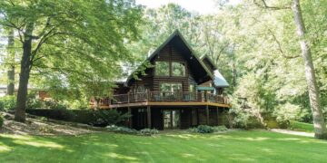 A large, multi-story log cabin with a steep gabled roof and expansive glass windows is nestled among mature, leafy green trees. A wide wooden deck with a dark railing wraps around the main level, overlooking a sprawling, neatly manicured green lawn. The lower level features glass doors leading out to a small stone patio. The scene is bright and airy, captured on a sunny day with soft shadows dappling the grass.