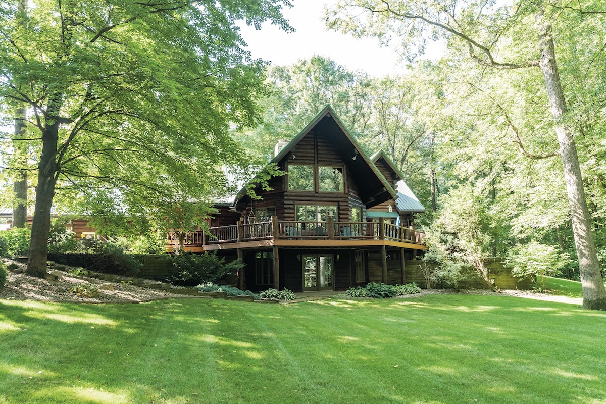 A large, multi-story log cabin with a steep gabled roof and expansive glass windows is nestled among mature, leafy green trees. A wide wooden deck with a dark railing wraps around the main level, overlooking a sprawling, neatly manicured green lawn. The lower level features glass doors leading out to a small stone patio. The scene is bright and airy, captured on a sunny day with soft shadows dappling the grass.
