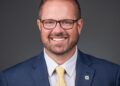 A professional studio headshot of Chris Jensen, the Mayor of Noblesville, Indiana. He is a man with short brown hair and brown-framed glasses, smiling broadly at the camera. He is dressed in a sharp navy blue blazer over a crisp white button-down shirt and a light yellow tie with subtle diagonal stripes. On his left lapel, he wears a small, square silver pin featuring a dark blue "N" logo, representing the Noblesville Millers. The background is a solid, neutral charcoal gray, which emphasizes his friendly and professional expression.