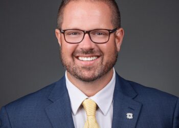 A professional studio headshot of Chris Jensen, the Mayor of Noblesville, Indiana. He is a man with short brown hair and brown-framed glasses, smiling broadly at the camera. He is dressed in a sharp navy blue blazer over a crisp white button-down shirt and a light yellow tie with subtle diagonal stripes. On his left lapel, he wears a small, square silver pin featuring a dark blue "N" logo, representing the Noblesville Millers. The background is a solid, neutral charcoal gray, which emphasizes his friendly and professional expression.