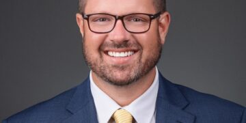 A professional studio headshot of Chris Jensen, the Mayor of Noblesville, Indiana. He is a man with short brown hair and brown-framed glasses, smiling broadly at the camera. He is dressed in a sharp navy blue blazer over a crisp white button-down shirt and a light yellow tie with subtle diagonal stripes. On his left lapel, he wears a small, square silver pin featuring a dark blue "N" logo, representing the Noblesville Millers. The background is a solid, neutral charcoal gray, which emphasizes his friendly and professional expression.