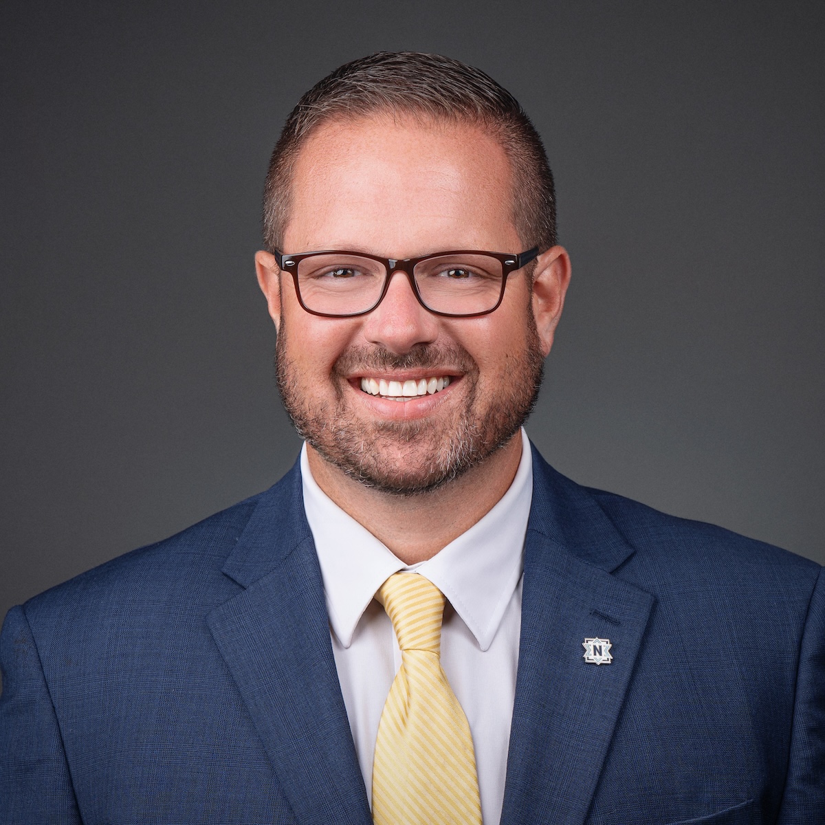 A professional studio headshot of Chris Jensen, the Mayor of Noblesville, Indiana. He is a man with short brown hair and brown-framed glasses, smiling broadly at the camera. He is dressed in a sharp navy blue blazer over a crisp white button-down shirt and a light yellow tie with subtle diagonal stripes. On his left lapel, he wears a small, square silver pin featuring a dark blue "N" logo, representing the Noblesville Millers. The background is a solid, neutral charcoal gray, which emphasizes his friendly and professional expression.