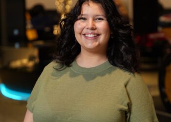A medium-shot, eye-level portrait of a smiling young woman with dark, shoulder-length curly hair. She is wearing a short-sleeved, olive green knit crew-neck top. She stands centered in the frame, facing the camera with a warm expression. The background is a modern, dimly lit interior, likely a museum or gallery, with a blue neon light glowing in the lower left and blurred metallic structures and warm lights behind her.