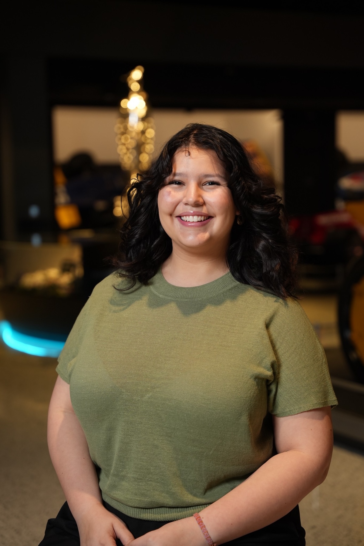 A medium-shot, eye-level portrait of a smiling young woman with dark, shoulder-length curly hair. She is wearing a short-sleeved, olive green knit crew-neck top. She stands centered in the frame, facing the camera with a warm expression. The background is a modern, dimly lit interior, likely a museum or gallery, with a blue neon light glowing in the lower left and blurred metallic structures and warm lights behind her.
