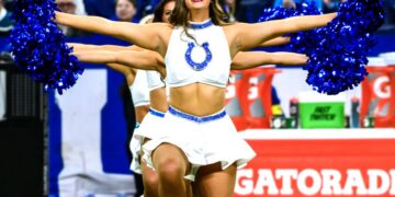 An Indianapolis Colts cheerleader smiles brightly while performing on the field. She wears a white sleeveless crop top with the blue horseshoe logo and a white flared skirt with blue rhinestone detailing at the waist. She holds large, metallic blue pom-poms out to her sides, with fellow cheerleaders lined up behind her in a synchronized pose. The background shows a crowded stadium and a red Gatorade cooler on the sideline.