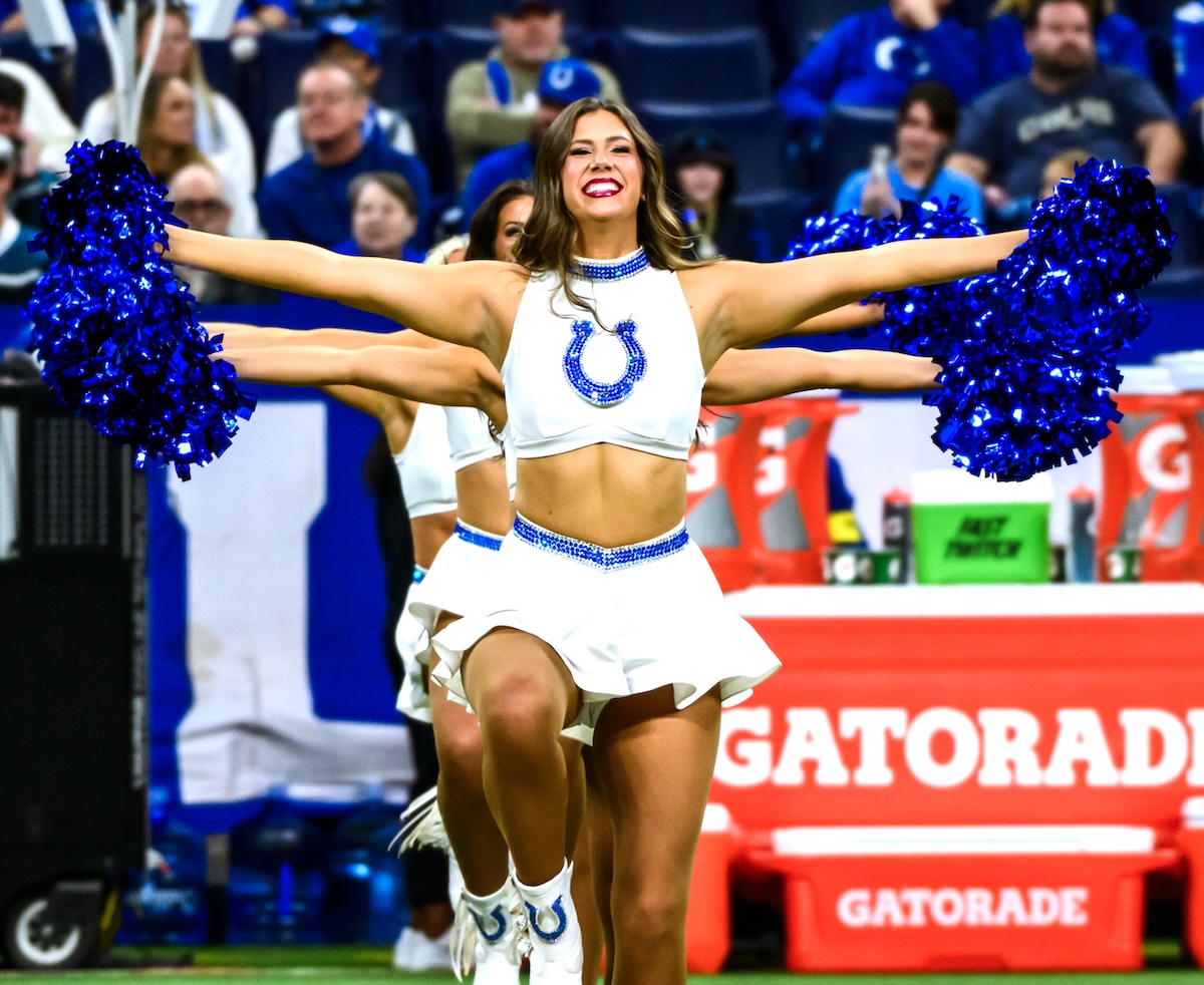 An Indianapolis Colts cheerleader smiles brightly while performing on the field. She wears a white sleeveless crop top with the blue horseshoe logo and a white flared skirt with blue rhinestone detailing at the waist. She holds large, metallic blue pom-poms out to her sides, with fellow cheerleaders lined up behind her in a synchronized pose. The background shows a crowded stadium and a red Gatorade cooler on the sideline.