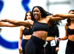 A young woman with long, dark curly hair and a joyful expression performs a dance routine in front of a blurred blue Indianapolis Colts horseshoe logo. She wears a black one-shoulder crop top and matching leggings, with a small identification tag pinned to her waist. Other dancers in similar black outfits are visible in the background, all mid-performance during an audition or rehearsal.