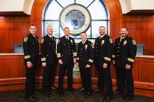 A formal group portrait of the Noblesville Police Department command staff posing inside the City Hall council chambers. Six officers stand in a row, dressed in their formal dark navy dress uniforms with gold braiding on their sleeves and badges pinned to their chests. They are positioned in front of a large, circular wooden seal of the City of Noblesville, which is mounted on a rich wood-paneled wall under an arched window. The team includes Chief Brad Arnold, Assistant Chief Eric Cunningham, and Deputy Chief Corey Everhart, representing the department's top leadership following their 2025 appointments.