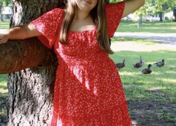 A young woman with long brown hair stands outdoors, leaning against a large tree trunk with her arms resting on low branches. She is smiling and wearing a short, red floral dress with flutter sleeves and brown cowboy boots. In the grassy background, several ducks are visible in the shade.
