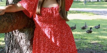 A young woman with long brown hair stands outdoors, leaning against a large tree trunk with her arms resting on low branches. She is smiling and wearing a short, red floral dress with flutter sleeves and brown cowboy boots. In the grassy background, several ducks are visible in the shade.