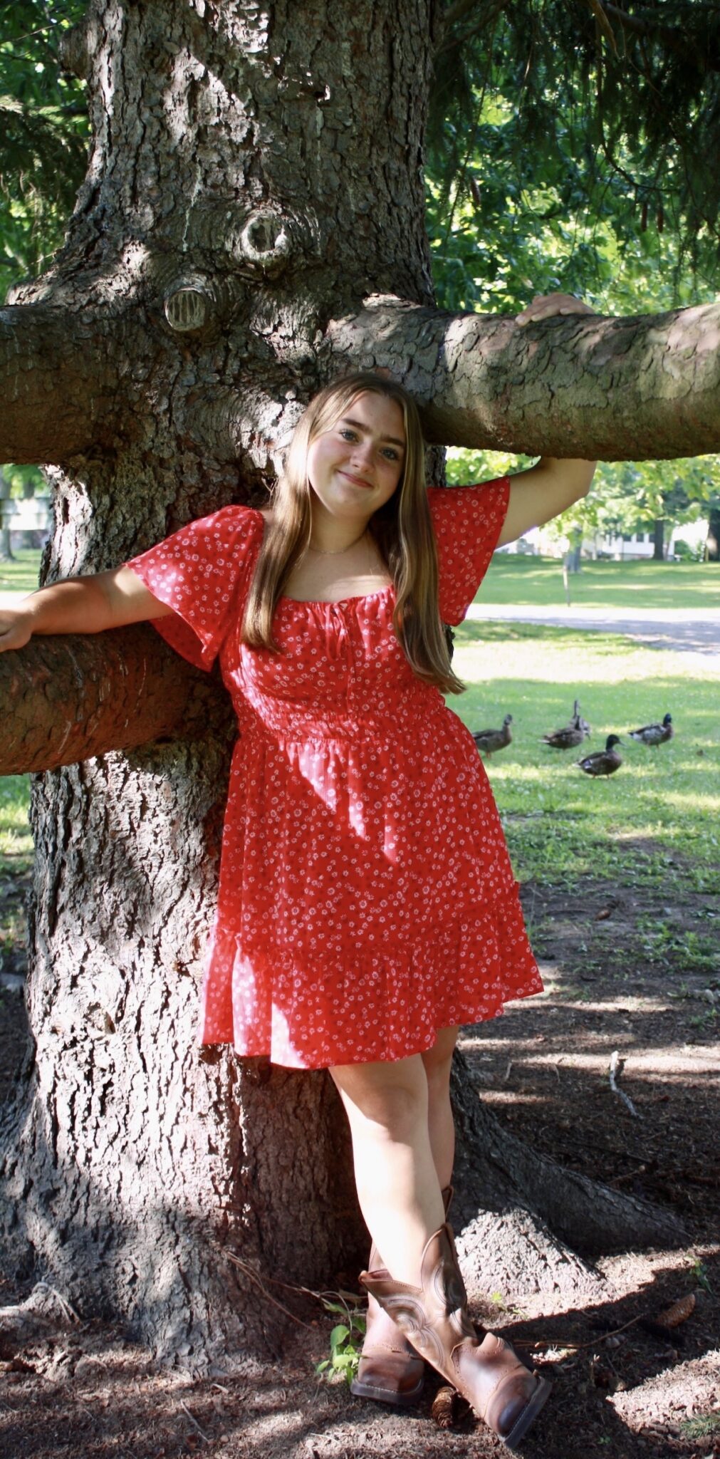 A young woman with long brown hair stands outdoors, leaning against a large tree trunk with her arms resting on low branches. She is smiling and wearing a short, red floral dress with flutter sleeves and brown cowboy boots. In the grassy background, several ducks are visible in the shade.