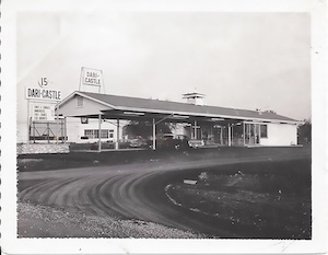 A black and white vintage photograph of an old-fashioned drive-in restaurant called "Dari-Castle." The building is a long, low structure with a flat roof and a large covered car-port area where several vintage cars are parked. A tall sign out front displays the restaurant's name and daily specials. The photo has a white border and the graininess of a classic film camera.