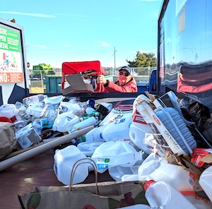 A first-person point-of-view shot looking into the bed of a pickup truck filled with various recyclables, primarily plastic milk jugs and white containers. In the background, a person in a red jacket and sunglasses is visible through the truck's rear window, holding up a red crate filled with more items to be sorted. A large green and white sign for a recycling center is partially visible on the left side of the truck bed.