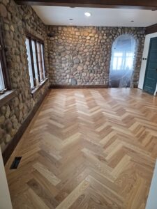 xAn empty room featuring a newly installed light oak herringbone-patterned hardwood floor. The walls are constructed from rustic, multi-colored fieldstone, and a large arched doorway on the right leads into a bright, sunlit space covered in protective plastic. Dark wood trim lines the windows and baseboards, contrasting with the light tones of the flooring.