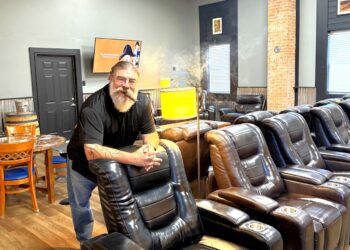 A medium full-shot of a man with a long, grey beard and glasses, identified as the owner of Hermanos Cigar Lounge in Noblesville, Indiana. He is wearing a black polo shirt and blue jeans, smiling while leaning against the back of a black power leather recliner. He has a lit cigar in his mouth, with a faint plume of smoke rising above his head. The lounge interior features several rows of similar black leather recliners with cup holders, a prominent brick pillar, grey walls, and a television screen in the background. A warm, yellow-shaded lamp stands next to him, and a small round table with blue chairs is visible to the left.