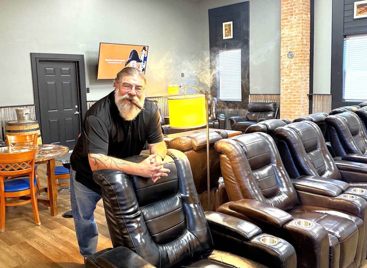 A medium full-shot of a man with a long, grey beard and glasses, identified as the owner of Hermanos Cigar Lounge in Noblesville, Indiana. He is wearing a black polo shirt and blue jeans, smiling while leaning against the back of a black power leather recliner. He has a lit cigar in his mouth, with a faint plume of smoke rising above his head. The lounge interior features several rows of similar black leather recliners with cup holders, a prominent brick pillar, grey walls, and a television screen in the background. A warm, yellow-shaded lamp stands next to him, and a small round table with blue chairs is visible to the left.