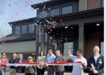 A celebratory outdoor scene at the ribbon-cutting ceremony for a modern, dark-colored home. A group of smiling people, including local officials and builders, stands on the front porch and walkway. A man in a blue shirt and blazer is in the center, cutting a wide red ribbon with ceremonial scissors while confetti rains down around them. In the foreground, a white tote bag with the text "Hope BLOOMS HERE" sits on the ground. The house features large windows and contemporary architecture, set against a bright blue sky.