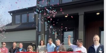 A celebratory outdoor scene at the ribbon-cutting ceremony for a modern, dark-colored home. A group of smiling people, including local officials and builders, stands on the front porch and walkway. A man in a blue shirt and blazer is in the center, cutting a wide red ribbon with ceremonial scissors while confetti rains down around them. In the foreground, a white tote bag with the text "Hope BLOOMS HERE" sits on the ground. The house features large windows and contemporary architecture, set against a bright blue sky.