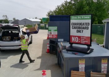 A bright outdoor scene at a recycling drop-off point. A person in a high-visibility yellow safety vest and a sun hat is unloading a large "Chewy" cardboard box from the back of a white SUV with its trunk open. They are walking toward a large blue industrial compactor. A prominent sign in the foreground reads "Clean, Dry, & Loose: CORRUGATED CARDBOARD ONLY," with a red "NO" section listing plastic, foam, strapping, and twine.
