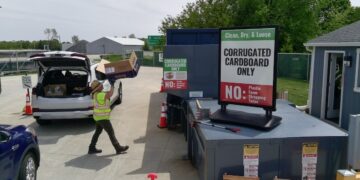 A bright outdoor scene at a recycling drop-off point. A person in a high-visibility yellow safety vest and a sun hat is unloading a large "Chewy" cardboard box from the back of a white SUV with its trunk open. They are walking toward a large blue industrial compactor. A prominent sign in the foreground reads "Clean, Dry, & Loose: CORRUGATED CARDBOARD ONLY," with a red "NO" section listing plastic, foam, strapping, and twine.