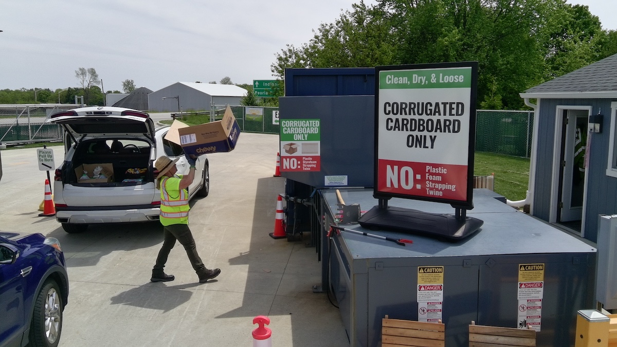 A bright outdoor scene at a recycling drop-off point. A person in a high-visibility yellow safety vest and a sun hat is unloading a large "Chewy" cardboard box from the back of a white SUV with its trunk open. They are walking toward a large blue industrial compactor. A prominent sign in the foreground reads "Clean, Dry, & Loose: CORRUGATED CARDBOARD ONLY," with a red "NO" section listing plastic, foam, strapping, and twine.