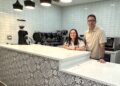 A man and woman stand behind a clean, modern cafe counter. The front of the counter is decorated with a black and white ornate tile pattern, topped with a smooth white countertop. Behind them is a white subway tile wall, a professional espresso machine, and a coffee grinder. Four stylish black pendant lights hang above the counter.