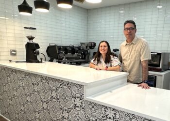 A man and woman stand behind a clean, modern cafe counter. The front of the counter is decorated with a black and white ornate tile pattern, topped with a smooth white countertop. Behind them is a white subway tile wall, a professional espresso machine, and a coffee grinder. Four stylish black pendant lights hang above the counter.