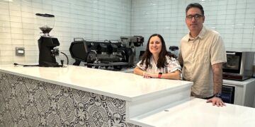 A man and woman stand behind a clean, modern cafe counter. The front of the counter is decorated with a black and white ornate tile pattern, topped with a smooth white countertop. Behind them is a white subway tile wall, a professional espresso machine, and a coffee grinder. Four stylish black pendant lights hang above the counter.