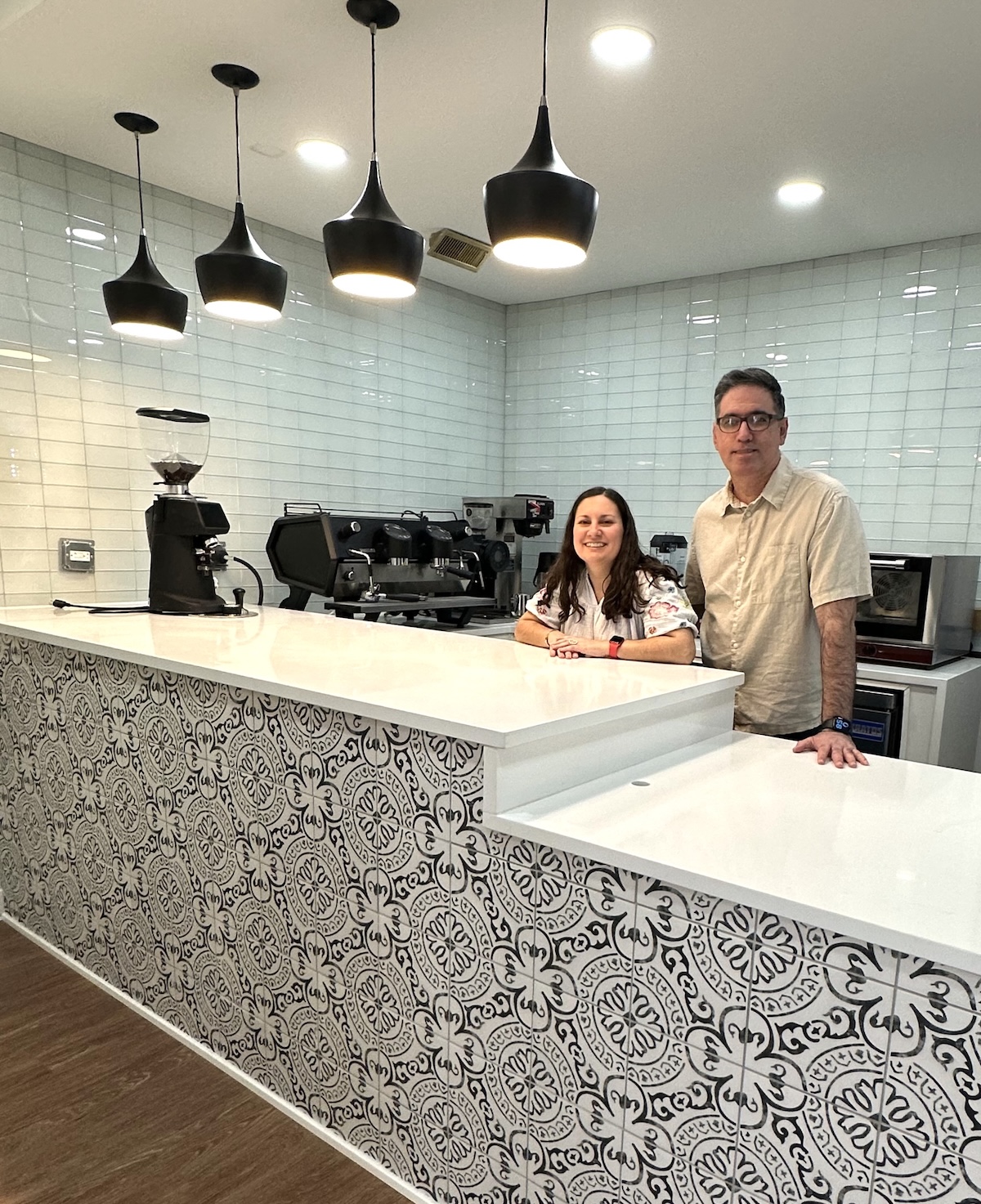 A man and woman stand behind a clean, modern cafe counter. The front of the counter is decorated with a black and white ornate tile pattern, topped with a smooth white countertop. Behind them is a white subway tile wall, a professional espresso machine, and a coffee grinder. Four stylish black pendant lights hang above the counter.