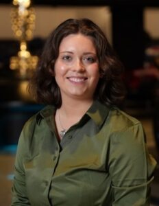 A professional headshot of a smiling young woman with shoulder-length, wavy brown hair. She is wearing an olive green, long-sleeved satin button-down shirt and a delicate silver necklace with a small heart-shaped pendant. The lighting is soft, creating a warm glow on her face, and the background is dark and out of focus, highlighting her as the central subject.