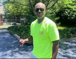 Medium shot of a man in a high-visibility neon yellow shirt with a small red and yellow logo, standing in a sunny outdoor setting with his arms at his sides.