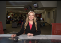 A medium shot of a smiling young woman, identified as Cameron Noe, an anchor and Managing Editor for NewsLink Indiana, sitting behind a sleek grey-and-black anchor desk. She is wearing a black blazer over a bright pink top and a gold necklace. The background is a busy, modern newsroom at Ball State University, with multiple computer workstations, journalists at their desks, and red office chairs. Large television monitors are mounted on the walls and ceiling, and the room is brightly lit with long, recessed LED light strips.