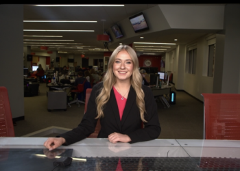 A medium shot of a smiling young woman, identified as Cameron Noe, an anchor and Managing Editor for NewsLink Indiana, sitting behind a sleek grey-and-black anchor desk. She is wearing a black blazer over a bright pink top and a gold necklace. The background is a busy, modern newsroom at Ball State University, with multiple computer workstations, journalists at their desks, and red office chairs. Large television monitors are mounted on the walls and ceiling, and the room is brightly lit with long, recessed LED light strips.