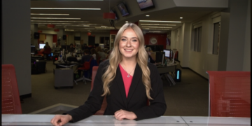 A medium shot of a smiling young woman, identified as Cameron Noe, an anchor and Managing Editor for NewsLink Indiana, sitting behind a sleek grey-and-black anchor desk. She is wearing a black blazer over a bright pink top and a gold necklace. The background is a busy, modern newsroom at Ball State University, with multiple computer workstations, journalists at their desks, and red office chairs. Large television monitors are mounted on the walls and ceiling, and the room is brightly lit with long, recessed LED light strips.