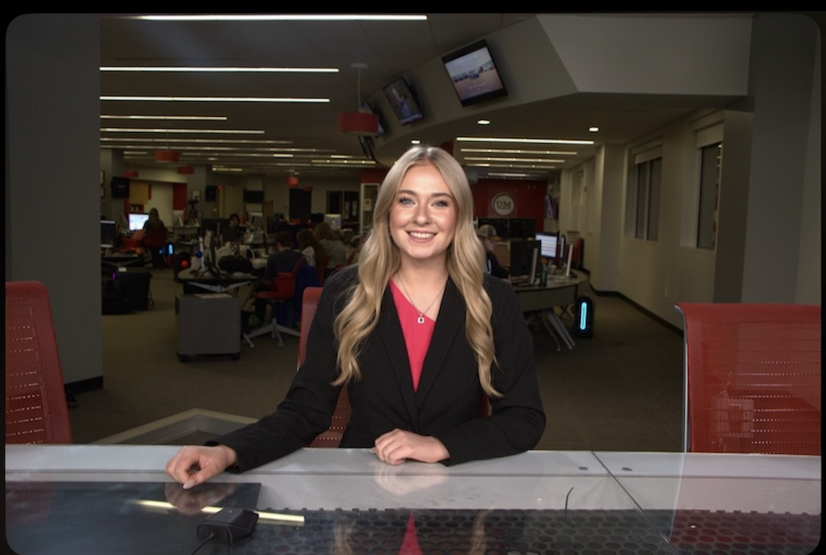 A medium shot of a smiling young woman, identified as Cameron Noe, an anchor and Managing Editor for NewsLink Indiana, sitting behind a sleek grey-and-black anchor desk. She is wearing a black blazer over a bright pink top and a gold necklace. The background is a busy, modern newsroom at Ball State University, with multiple computer workstations, journalists at their desks, and red office chairs. Large television monitors are mounted on the walls and ceiling, and the room is brightly lit with long, recessed LED light strips.
