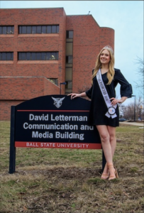 A full-length, eye-level shot of a smiling young woman, identified as Cameron Noe, a 2026 500 Festival Princess, posing next to the entrance sign for the David Letterman Communication and Media Building at Ball State University. She is wearing a black long-sleeved mini dress with bell sleeves, tan block-heeled sandals, a small sparkling tiara, and a white sash that reads "500 FESTIVAL PRINCESS 2026." She has one hand on the black-and-red sign and the other on her hip. Behind her is a large, multi-story brick university building under a bright, overcast sky.