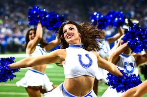 n Indianapolis Colts cheerleader with brown hair and red lipstick performs on a football field at night. She is smiling and looking off-camera, wearing the team's signature white sleeveless crop top with the blue horseshoe logo and a matching white flared skirt. She holds bright blue pom-poms with her arms extended, while other cheerleaders in the same uniform are visible behind her, blurring into the stadium lights.