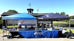 An outdoor event setup featuring a long table covered with a blue tablecloth under two pop-up canopies—one blue and one black. Six people are seated behind the table, which holds various flyers and informational signs. The blue tablecloth prominently displays the name "St. Matthews Area Ministries" in white text. The scene is set on a sunny day in front of a dark building with a cupola and weather vane, likely at a golf course or community park.