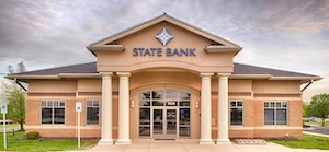 The exterior of the State Bank Zionsville branch, a single-story brick building with a grand entrance featuring four tall, cream-colored columns supporting a portico. The "STATE BANK" name and a blue diamond logo are mounted on the facade above the doors. The sky is overcast with soft lighting, and the building is flanked by green trees and a parking lot.