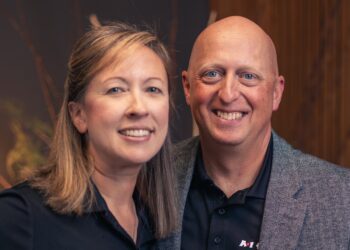 A professional portrait of a man and a woman standing close together and smiling. Both are wearing black polo shirts with a small white "A1" logo on the chest. The man, on the right, is bald and wearing a grey textured blazer over his polo. The woman, on the left, has shoulder-length light brown hair. They are posed against a warm, out-of-focus background that features dark wood paneling and decorative natural branches.